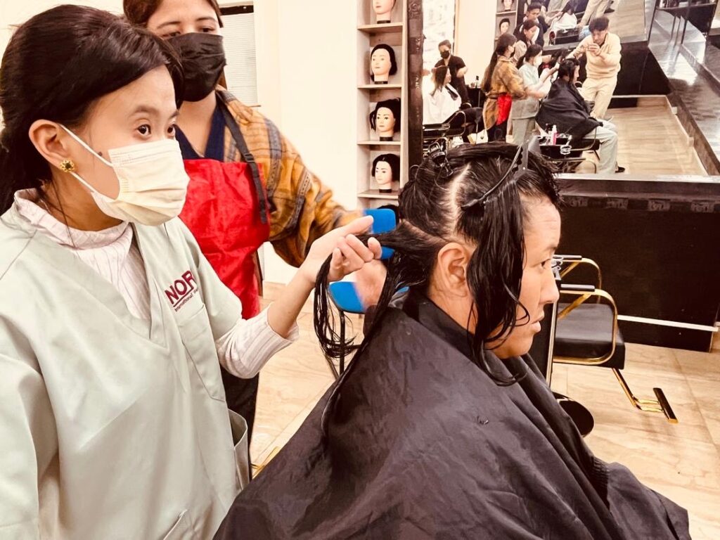 A trainee wearing a face mask and a light green uniform is combing a seated client's wet hair, sectioned and clipped, in a modern salon with mirrors and other clients in the background.