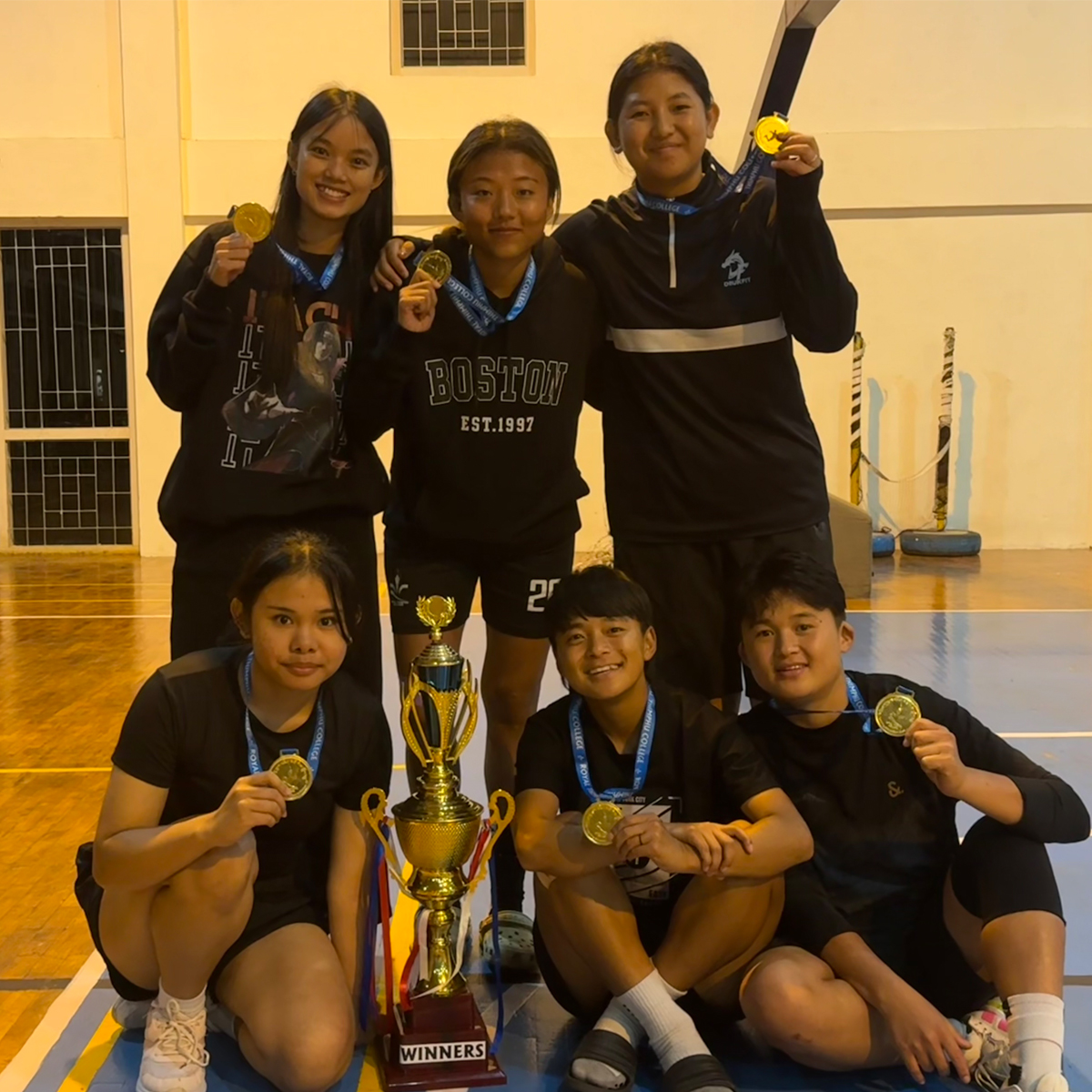 Six basketball players, wearing medals, pose with a large trophy labeled "Winners" on an indoor court.