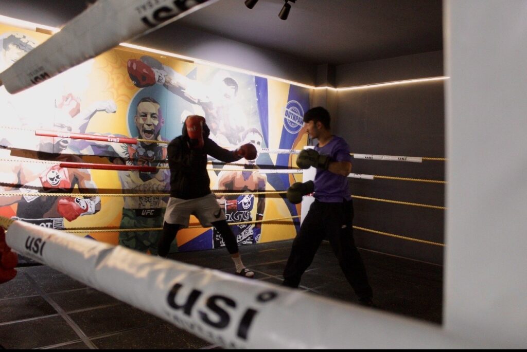 Two people sparring inside a boxing ring wearing gloves and protective gear, with a colorful mural of fighters on the wall in the background