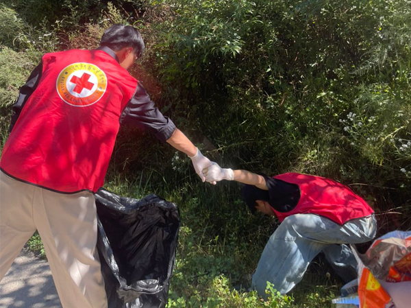 Two Bhutan Red Cross Society RTC Club volunteers clean up roadside waste, with one reaching into the bushes while the other assists and holds a garbage bag.