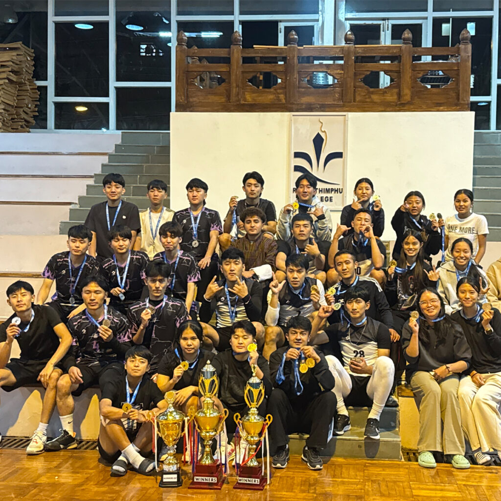 Group of young men and women in sportswear and casual clothes sit and stand on indoor bleachers, holding medals and posing behind several large trophies, with the "Thimphu College" logo displayed on the wall.
