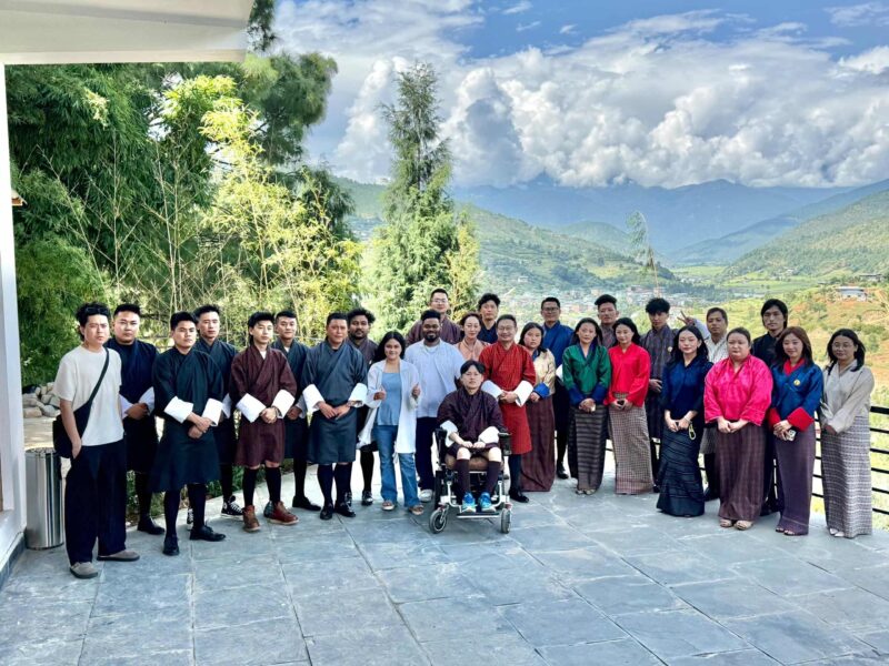 Group photo of Bhutanese artists, including a person in a wheelchair, taken during a vocal workshop in Punakha, Bhutan, with scenic mountains in the background.