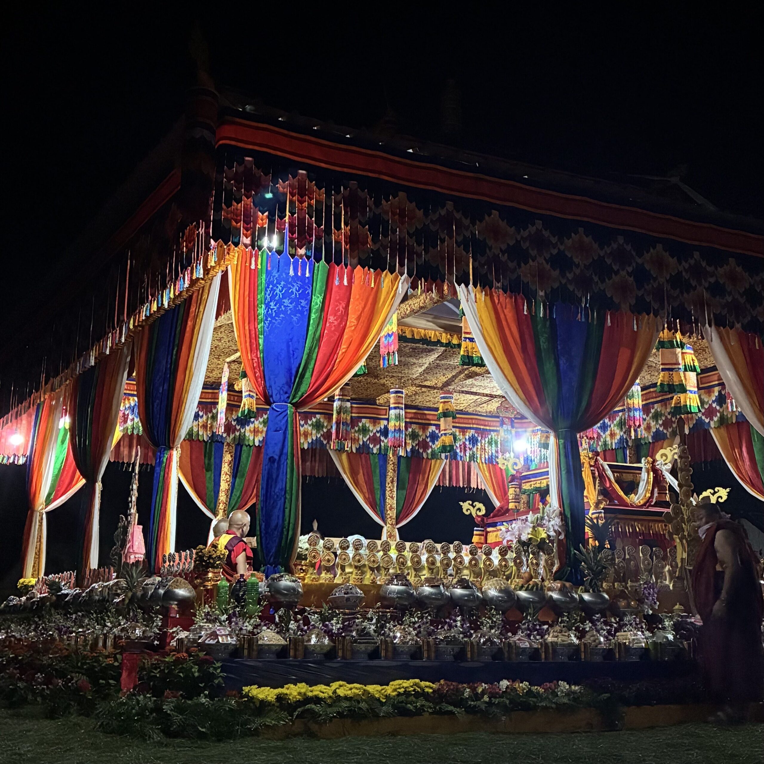 A beautifully decorated ceremonial canopy adorned with vibrant colorful fabrics and lights, set against a dark night sky, showcasing offerings arranged in front for a state funeral ceremony.
