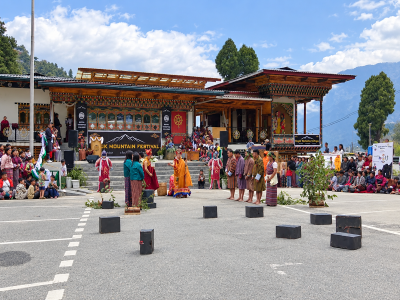 People in traditional Bhutanese clothing gather in front of an ornate Bhutanese building during a cultural festival, with musical equipment and black speakers arranged in the courtyard.